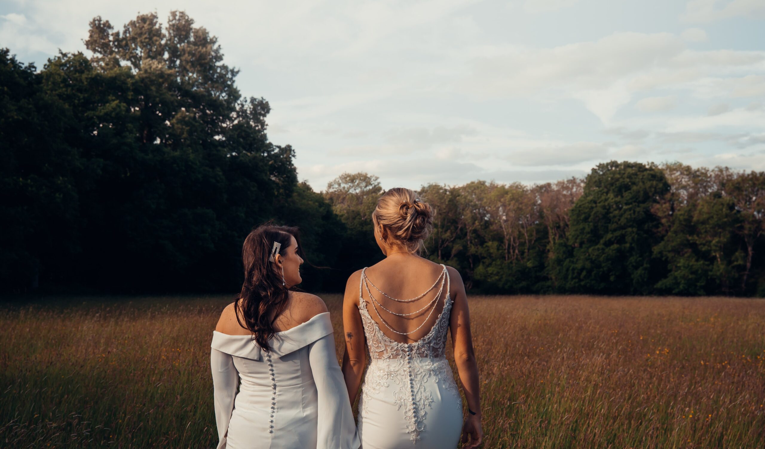 Two brides walking away through a field at dusk, a joyful same sex wedding celebration